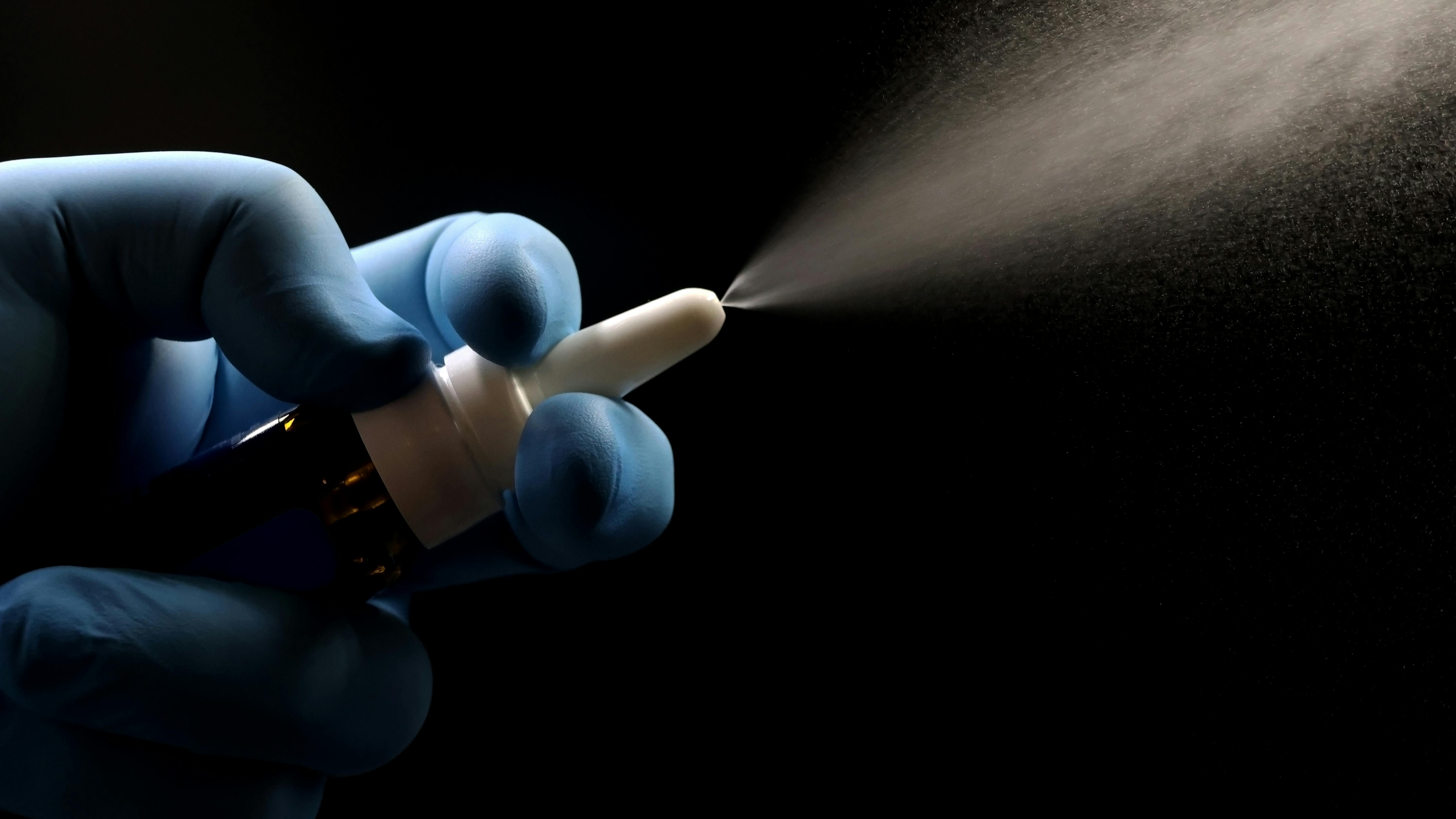 A gloved hand holding a nasal spray bottle, actively dispensing a fine mist spray against a black background.