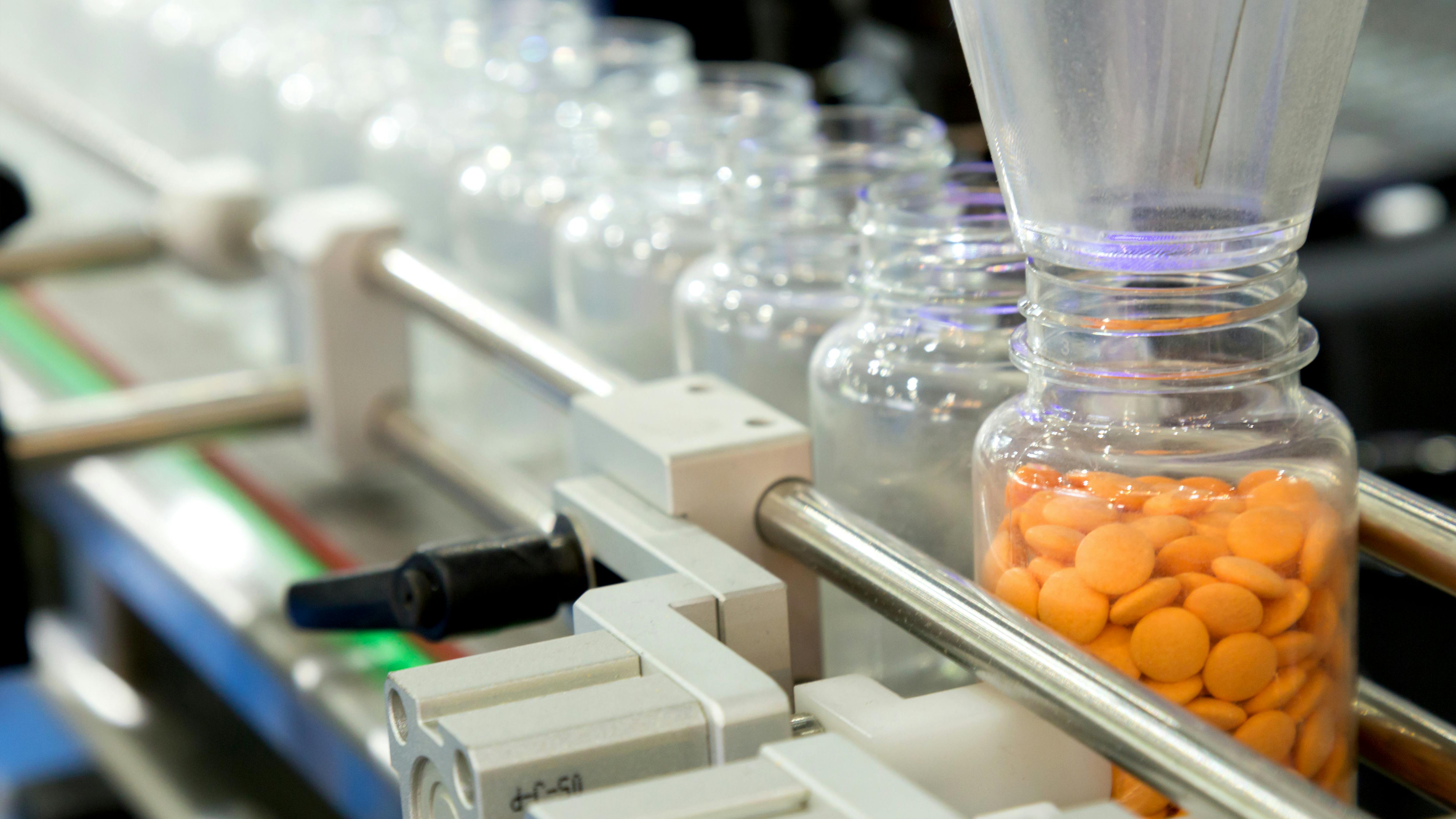 Pharmaceutical manufacturing equipment dispensing orange tablets into a clear glass bottle.