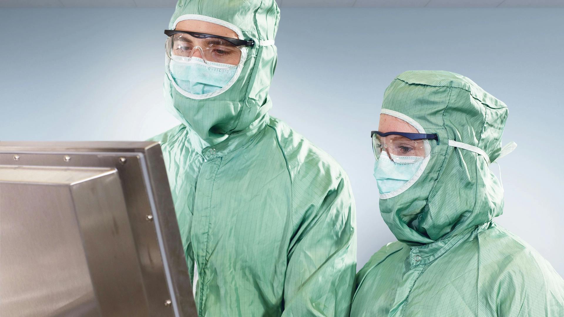 Two cleanroom workers in green protective suits and masks review data on an industrial monitor.