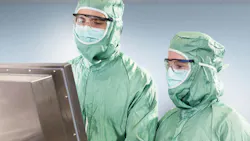 Two cleanroom workers in green protective suits and masks review data on an industrial monitor. Two cleanroom workers in green protective suits and masks review data on an industrial monitor.
