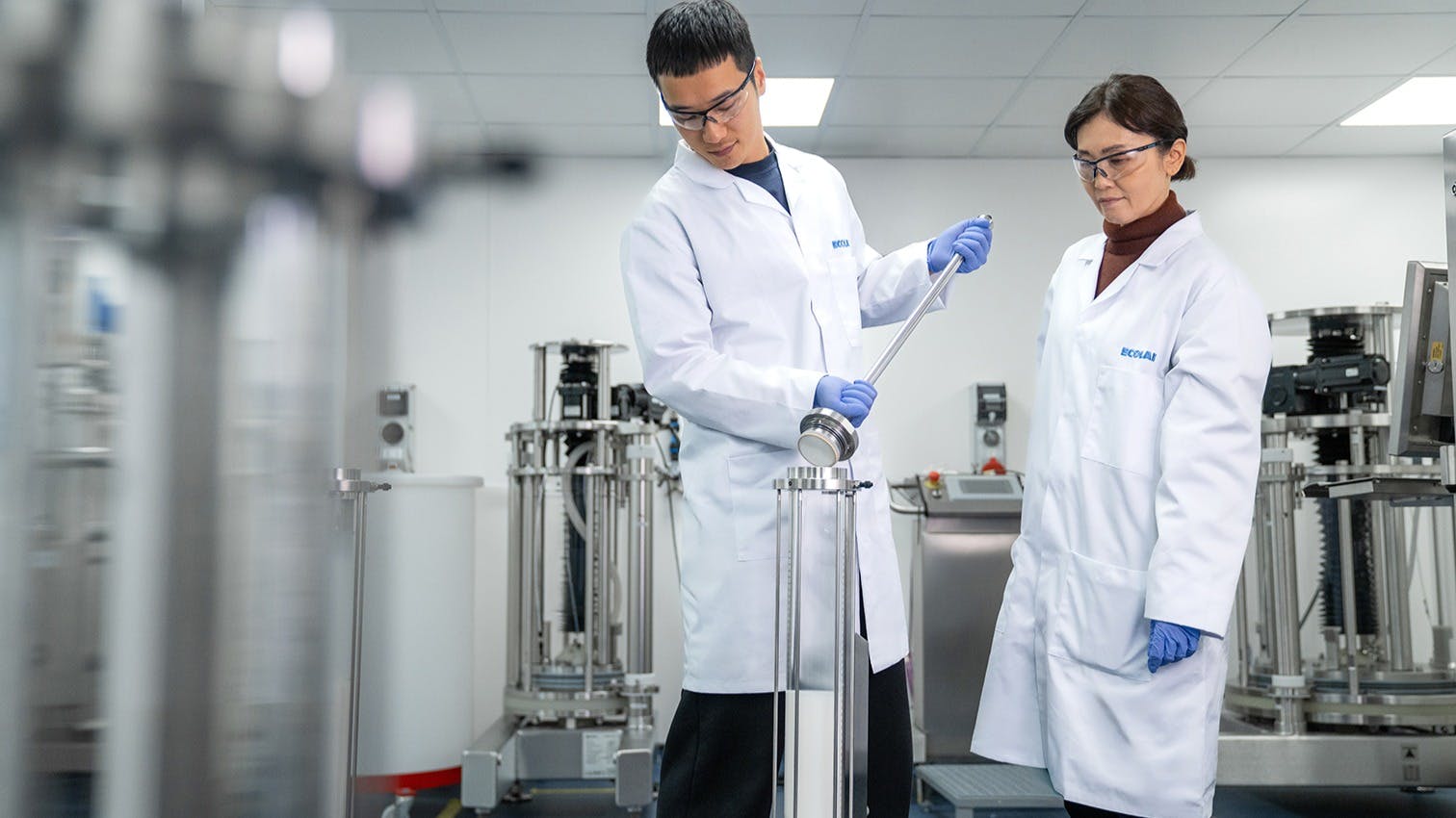 Two Ecolab scientists in white lab coats and blue gloves examine equipment in a modern laboratory.