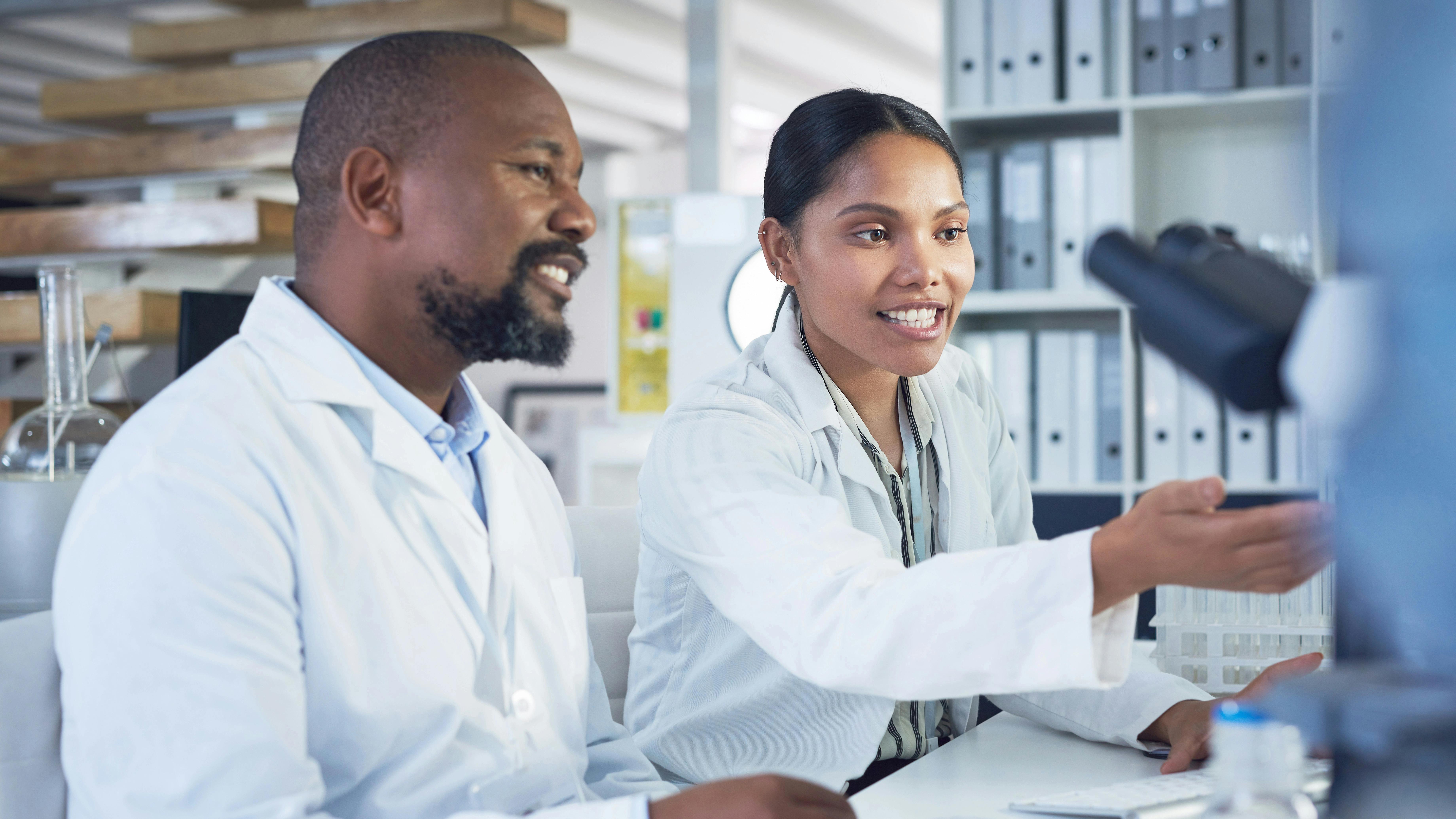 Two smiling scientists in white lab coats collaborate at a laboratory workstation with a microscope nearby.
