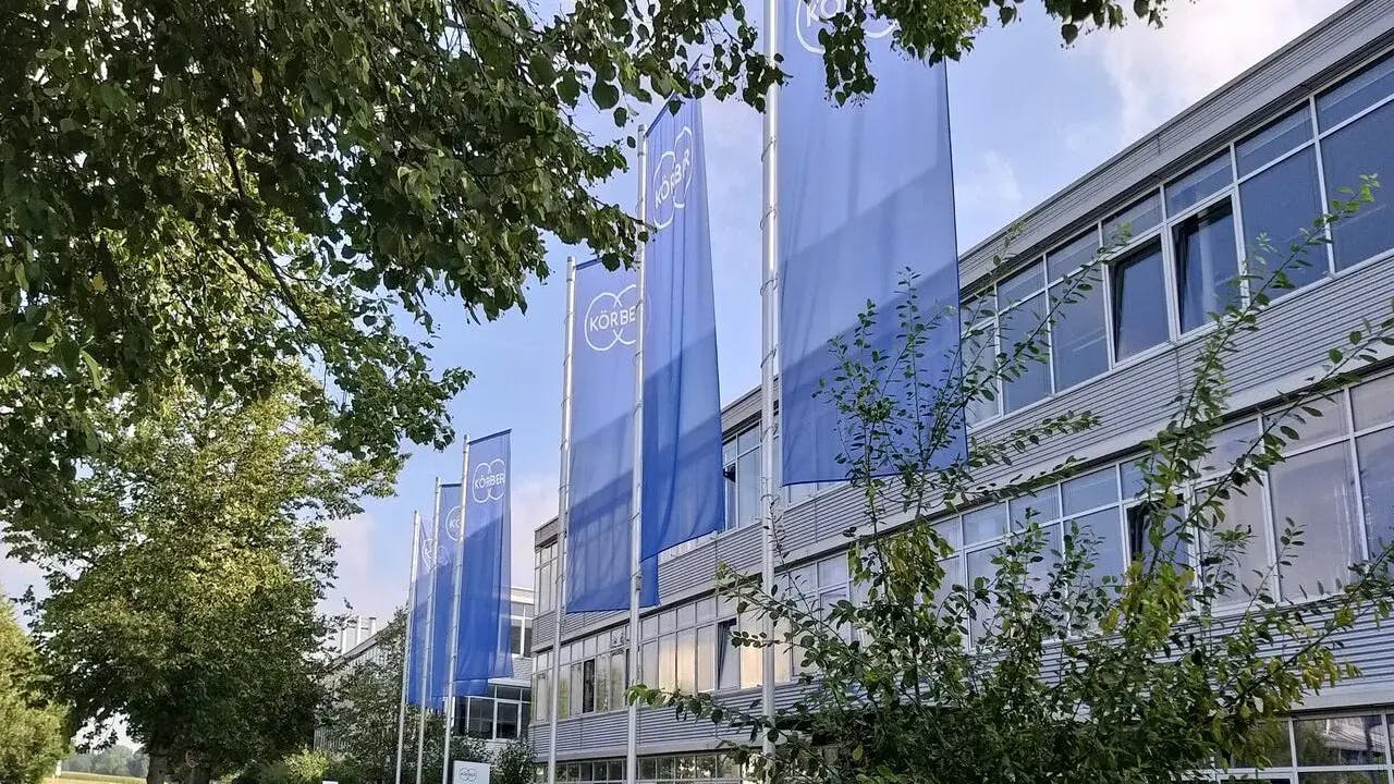 K&ouml;rber flags fly in front of corporate building surrounded by trees in foreground