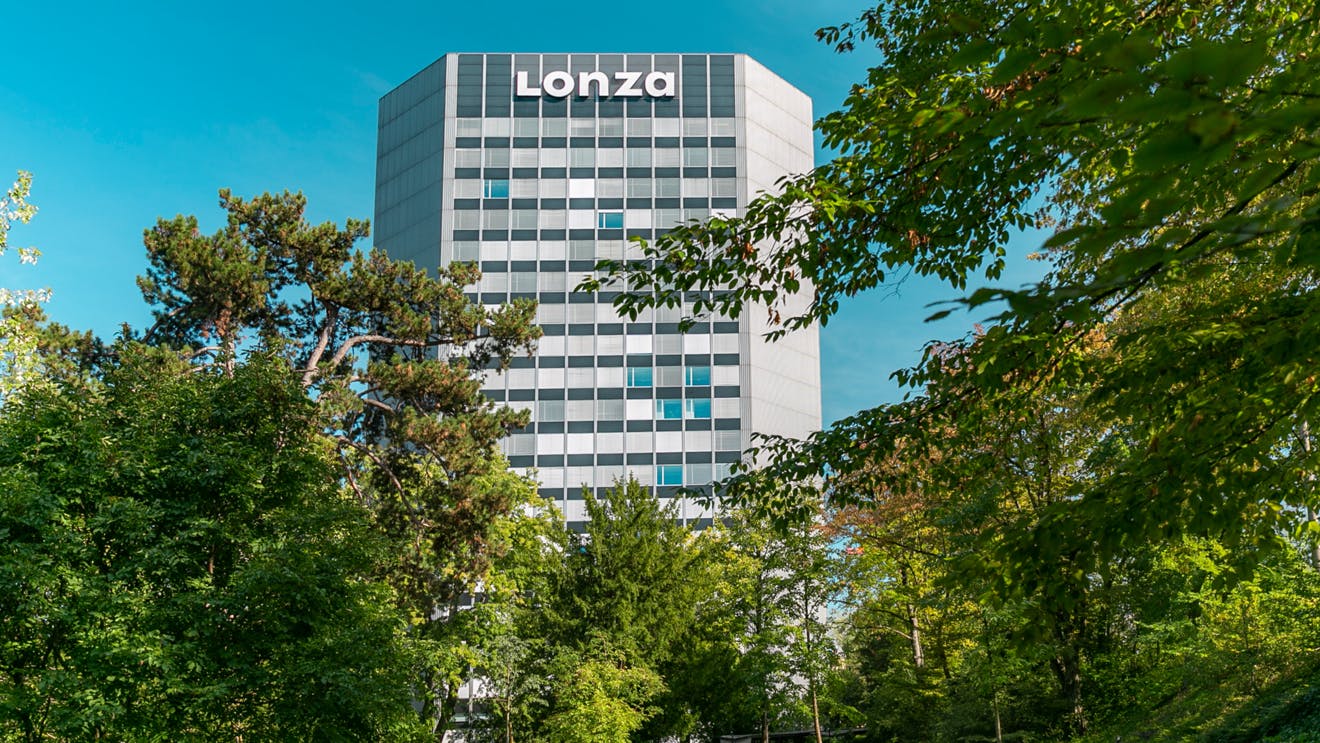 Lonza's corporate headquarters tower in Basel, Switzerland as seen through treeline on bright sunny day