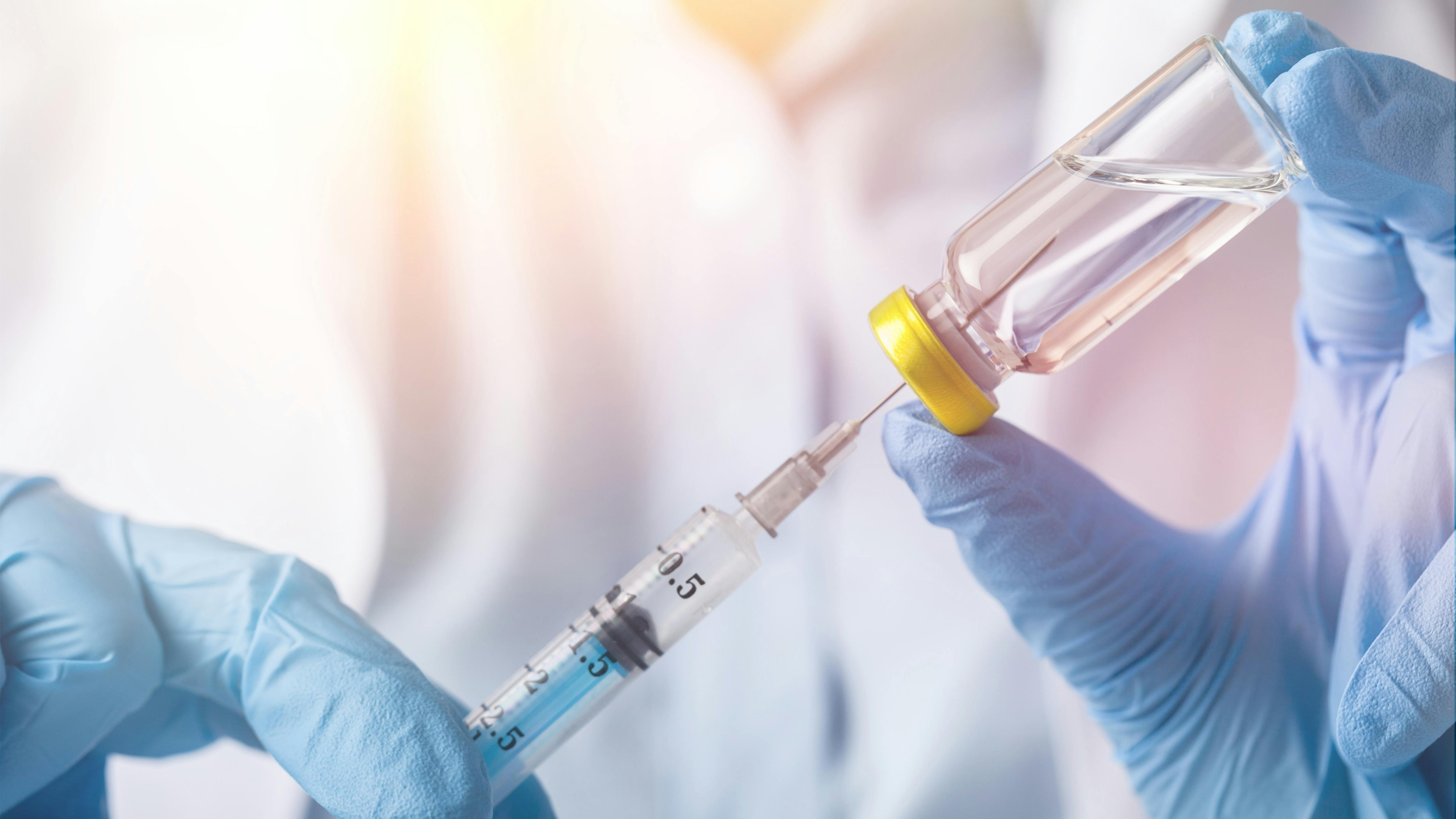 Gloved hands draw liquid from a glass medication vial into a syringe in a clinical setting.