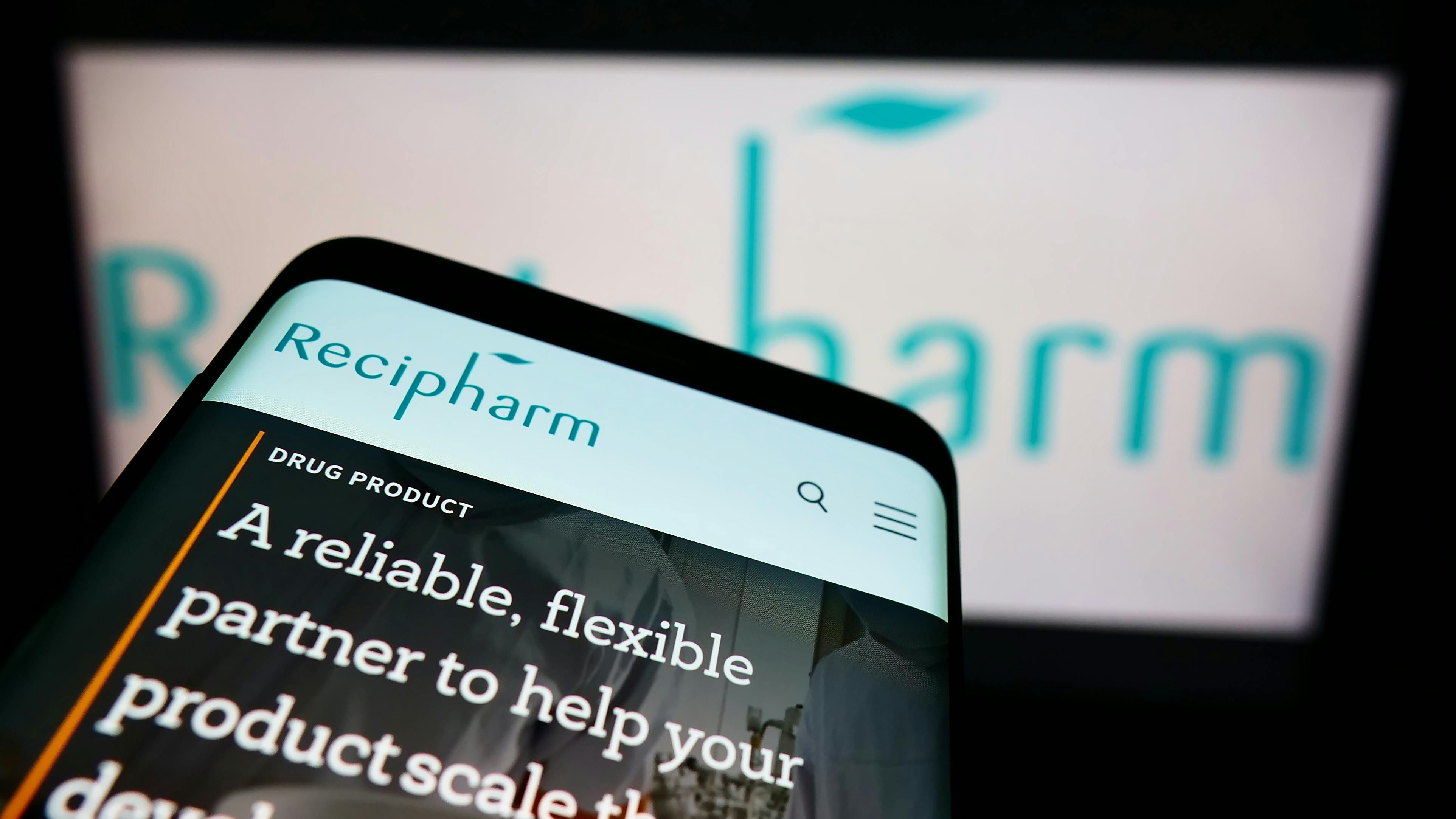 Recipharm website displayed on smartphone with logo blurred on digital screen in background