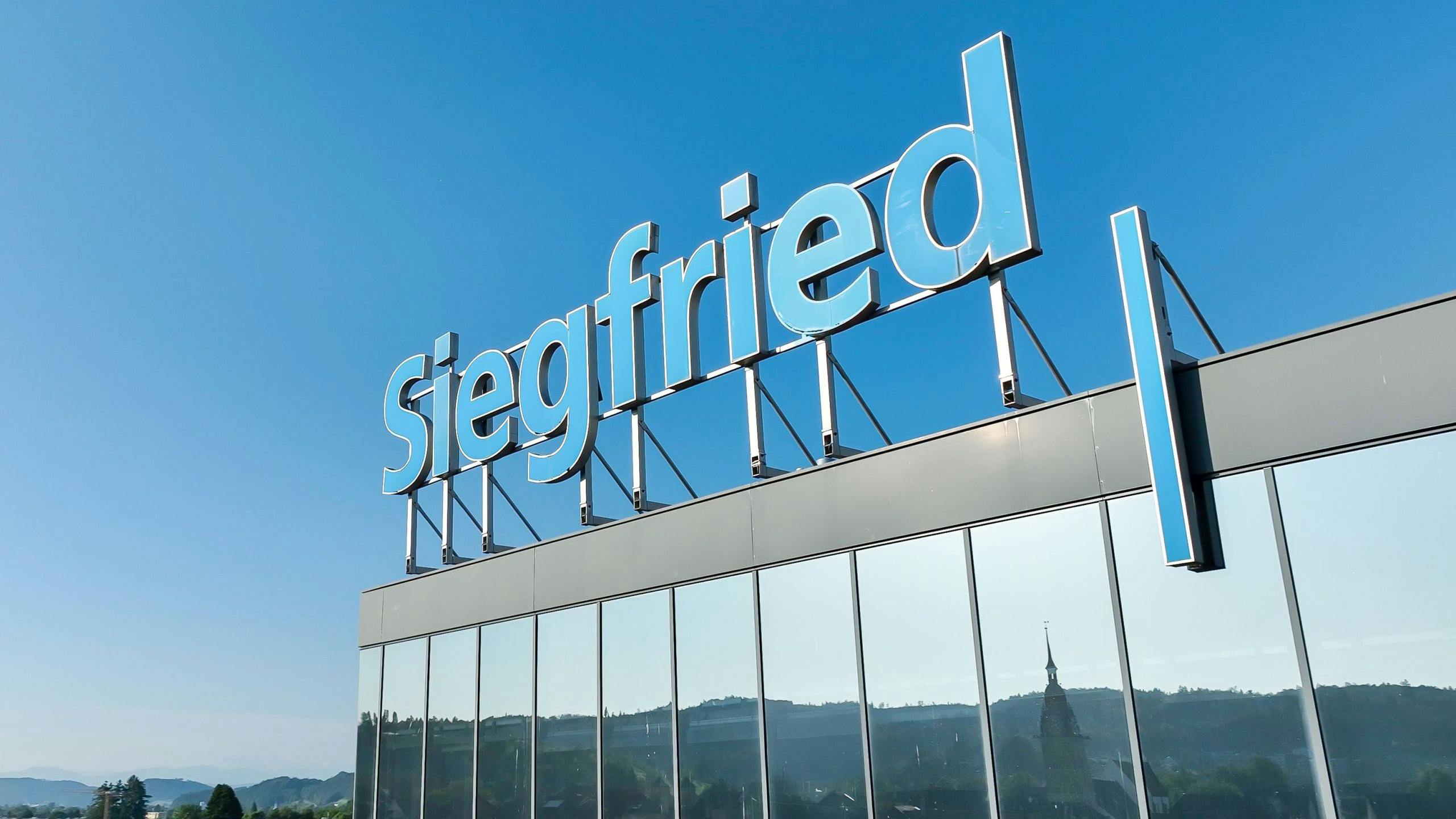 Siegfried sign on top of headquarters building against a blue sky. Landscape reflects in the building windows.