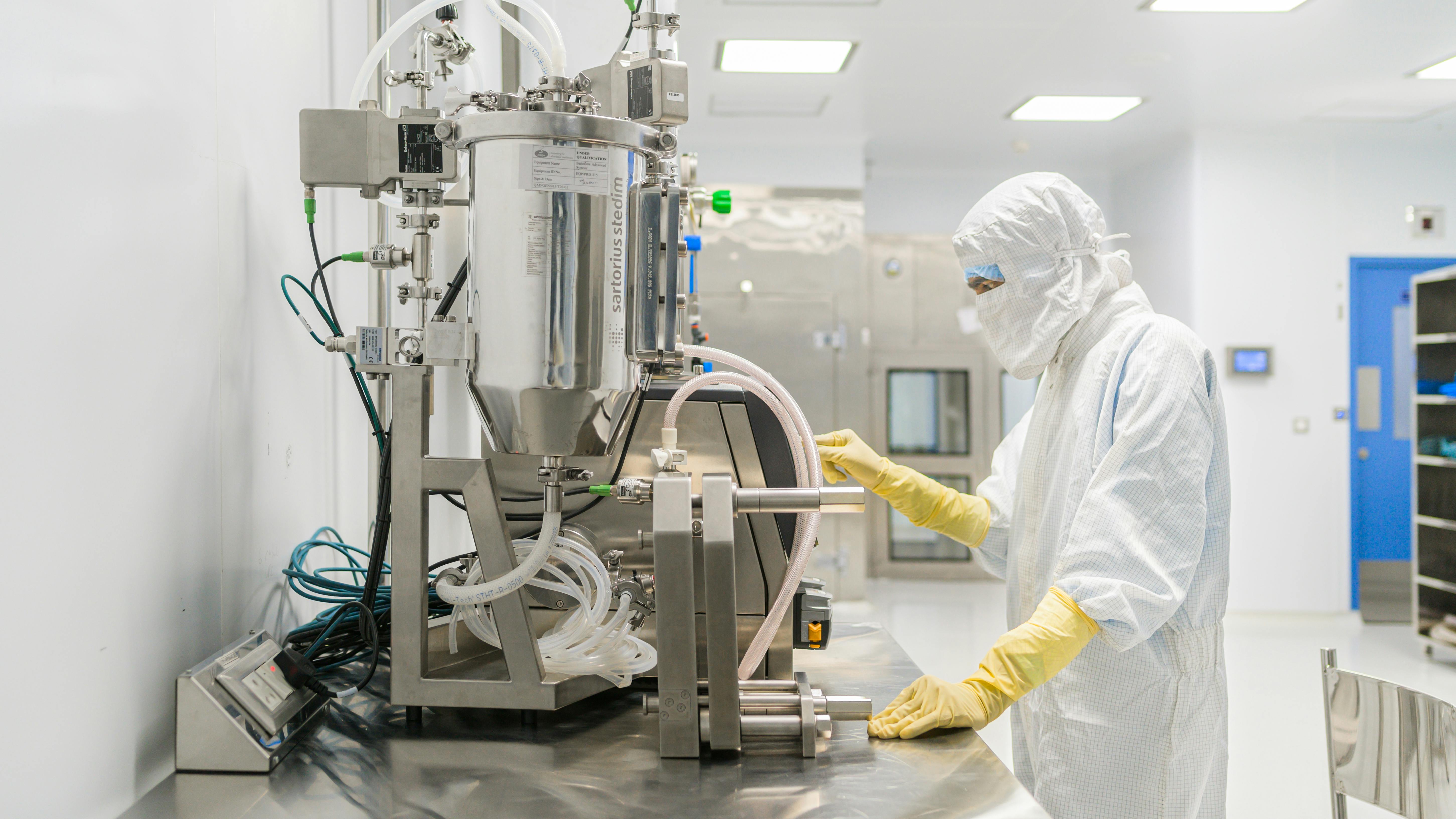 Cleanroom technician in protective gown and gloves operating stainless steel bioprocessing equipment in a pharmaceutical manufacturing facility.