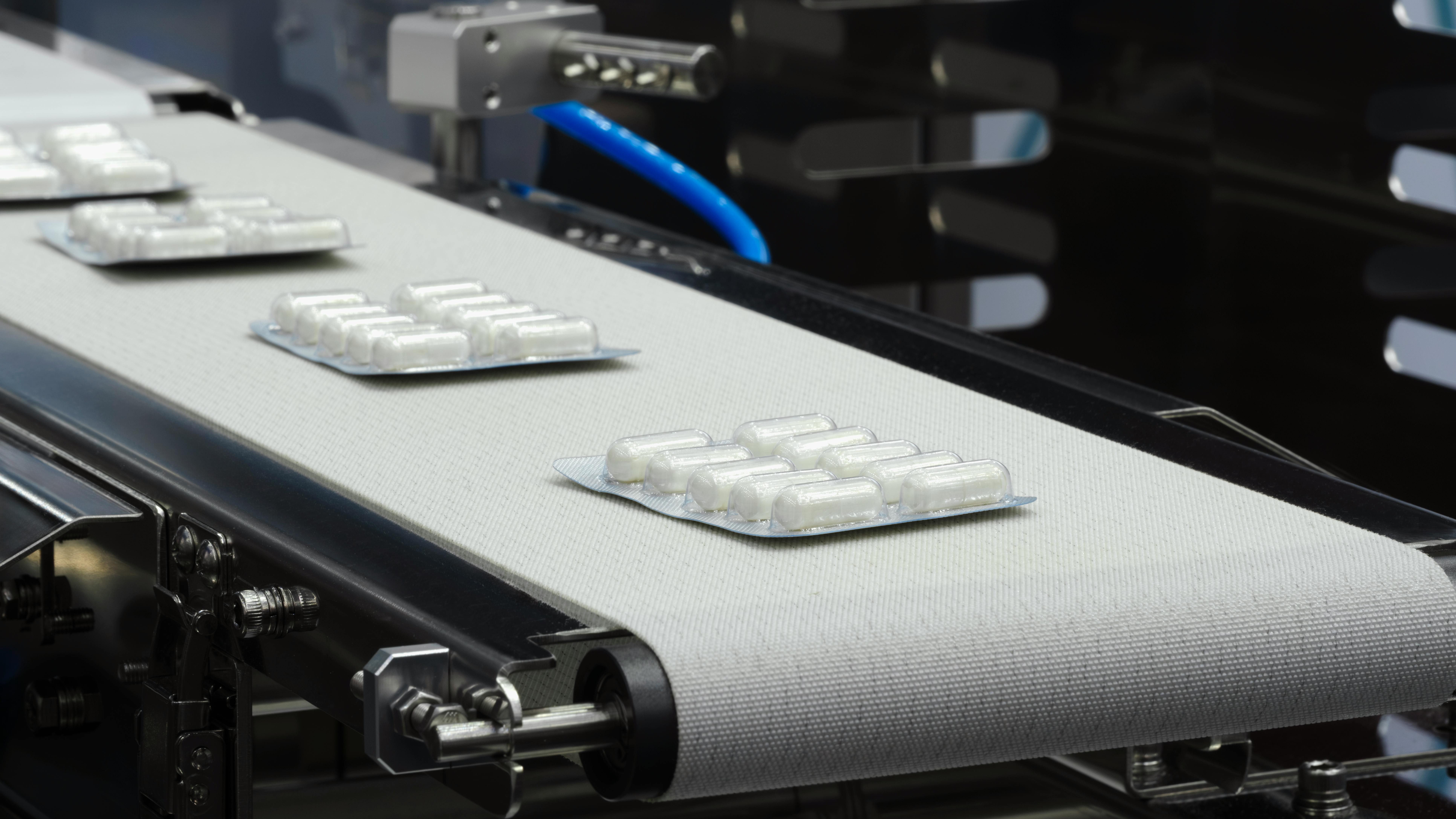 Blister packs of white capsules moving along a conveyor belt on a pharmaceutical manufacturing line, illustrating automated drug packaging and production.