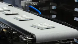 Blister packs of white capsules moving along a conveyor belt on a pharmaceutical manufacturing line, illustrating automated drug packaging and production.